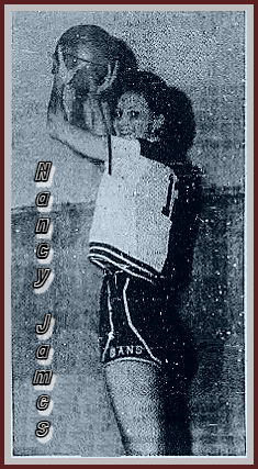 Side view of Nancy James, Gans High School basketball player holding up a basketball in front of her but above her head, looking bck at camera, GANS in white on bottom of side of her shrots. From the Sequoyah County Times, Sallislaw, Oklahoma, February 23, 1967.