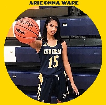 Picture of Arieonna Ware, girls high school player for Central High School in Battle Creek, Michigan. In black uniform with yellow lettering, number 15, in front of folded stands, holding basketball towards us with one arm, her right hand holding the ball.