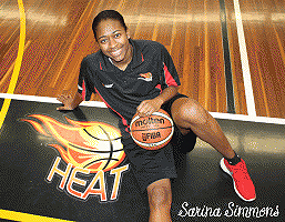 Sarina Simmons, Midura LAdy Heat basketbaall player, sitting on court, with basketball, next to Heat logo.